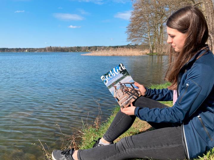 Eine Frau hält sitzend am Ufer das Gästemagazin Seeblick in der Hand © Seenland Oder-Spree