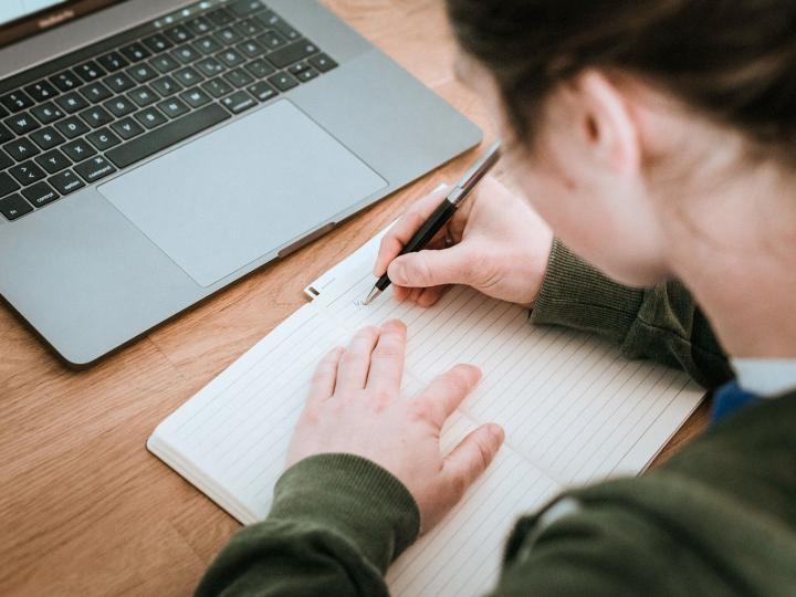 Eine junge Frau schreibt Notizen in ein Heft vor einem Laptop © Hagar Lotte Geyer/Pixabay